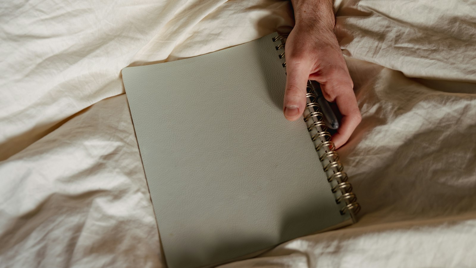 A hand holding a closed notebook and a pen on rumpled bed sheets
